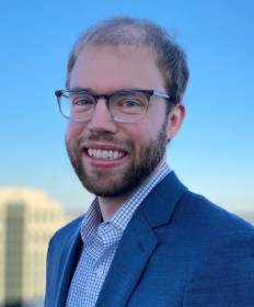 Lucas Henneman wears a dark blue suit and glasses for his faculty profile in the Department of Civil, Environmental, and Infrastructure Engineering.